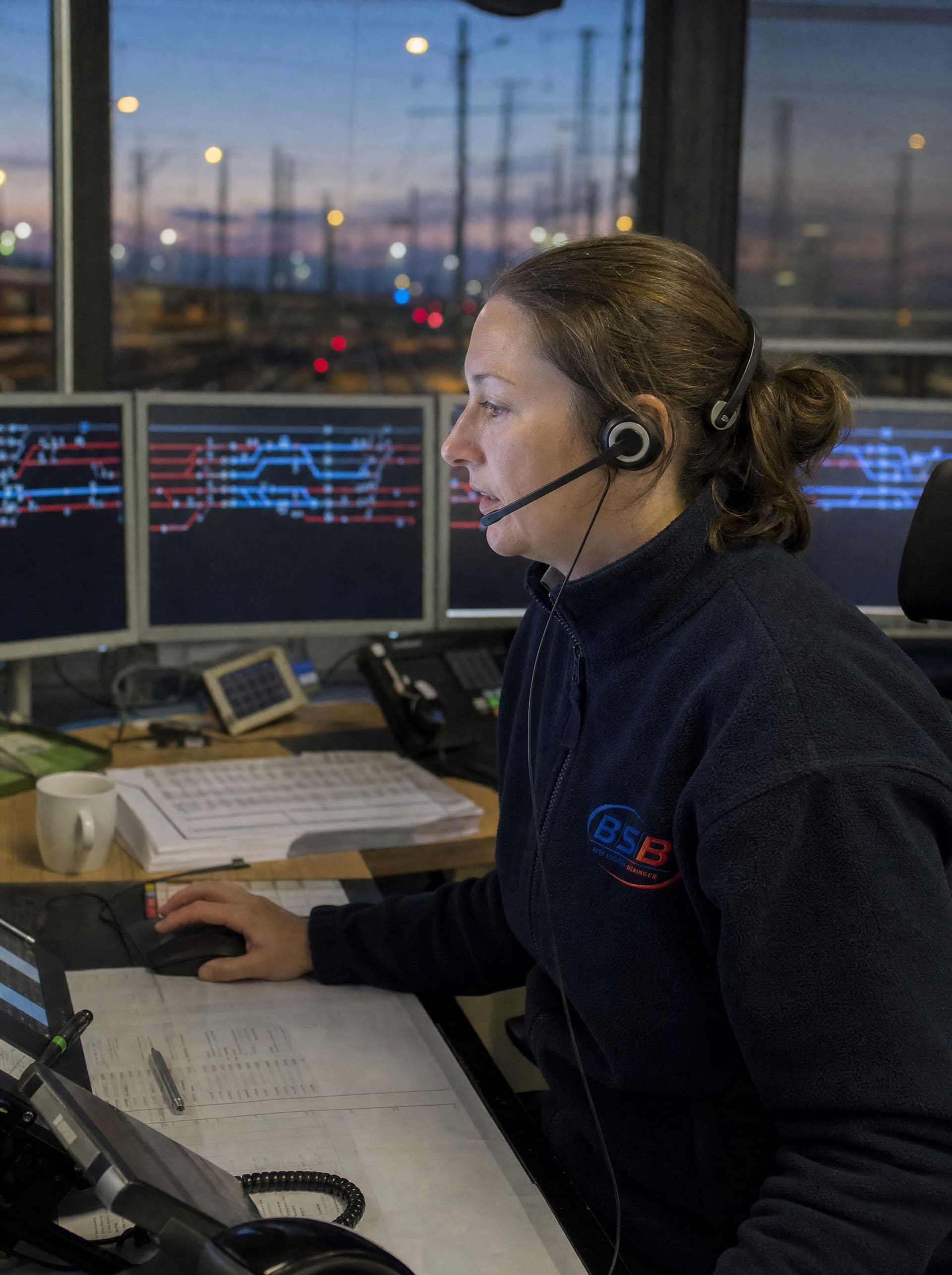 Disponentin mit Headset in BSB-Fleecejacke im Betriebszentrum, dahinter ein Bahnhof in der Abenddämmerung